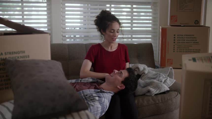 Woman and man sit in on a couch surrounded by moving boxes