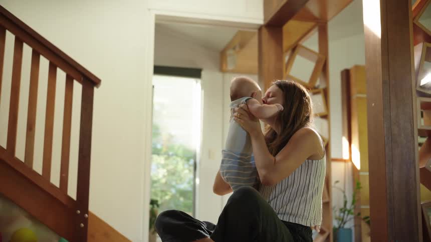 A joyful woman sits indoors lifting a baby above her head while smiling warmly