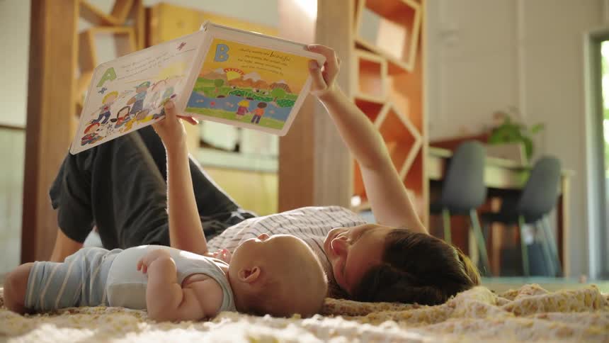 Mother lying on the living room rug reading a book with her baby next to her