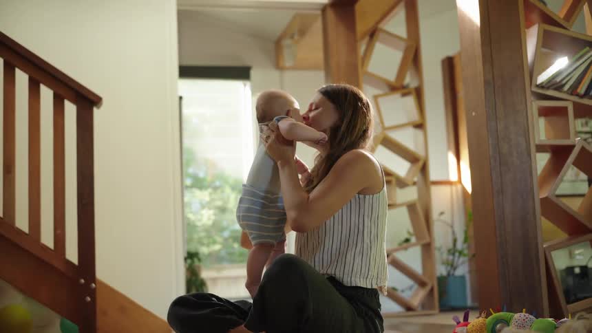 A woman sitting on the floor near a staircase in her living room at midday lifting her baby up while playing and laughing together