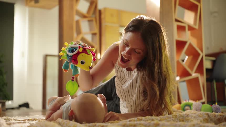 A mother plays with her baby who is lying on a blanket using a colorful plush insect toy