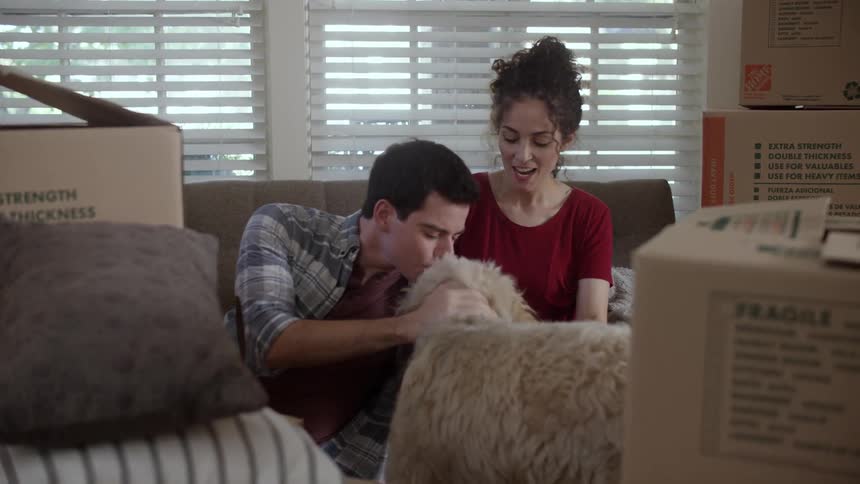 A man and woman pet a dog with cardboard boxes surrounding them in a living room