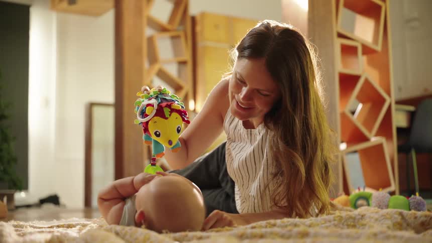 A woman lovingly interacts with her baby showing them a colorful toy while they lie on a play mat