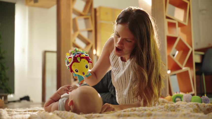 A woman playfully holds a toy above a babys face as the baby lies on the bed