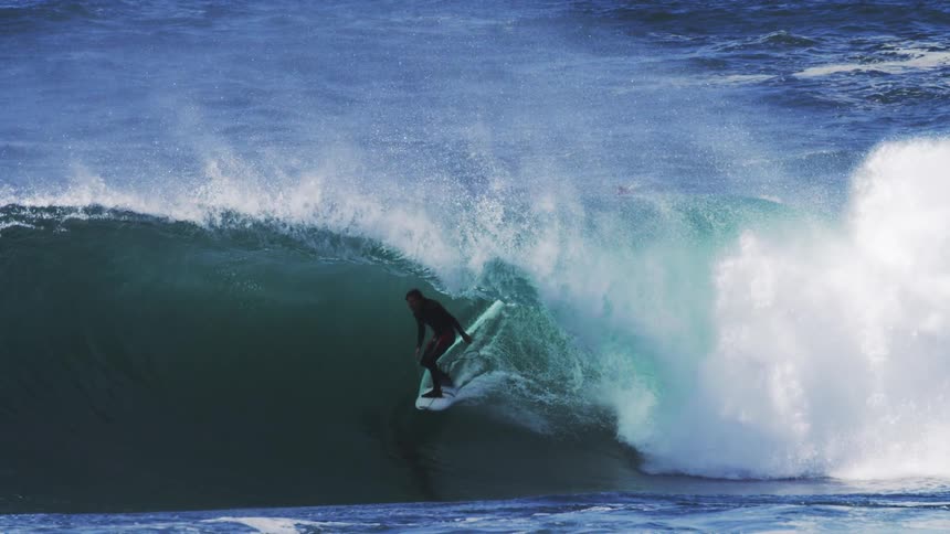 A surfer silhouetted against a powerful blue wave appears to be riding it skillfully with spray and foam erupting around them