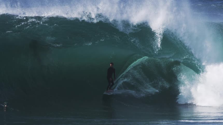 A surfer rides a powerful dark blue wave thats about to break with another figure visible further down the line