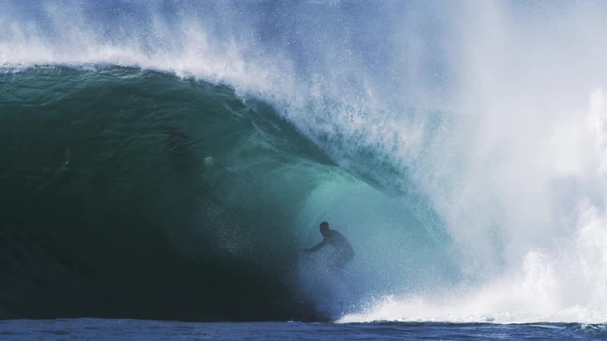 A surfer emerges from beneath a towering wave skillfully riding through the ocean under the midday sun