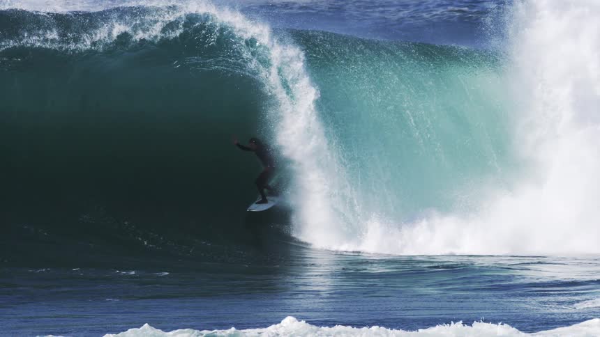 A surfer carves down the face of a massive turquoise wave under a moody sky