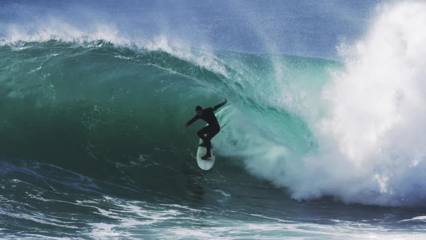A surfer skillfully rides deep inside the barrel of a large powerfully curling ocean wave in slow motion with immense spray jetting from the breaking crest