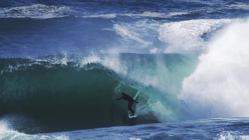 A surfer rides a massive wave in the ocean under the bright midday sun
