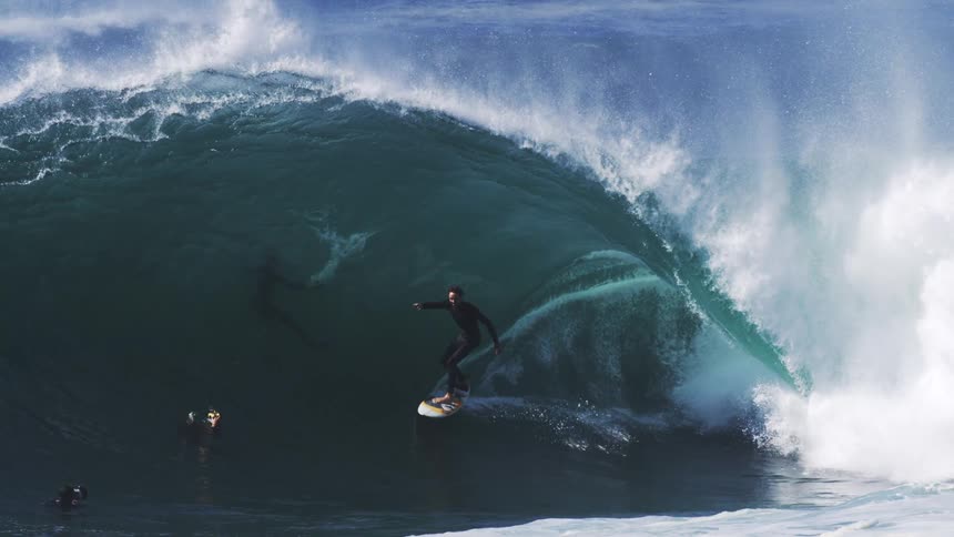 A person rides a gigantic ocean wave on a surfboard with two other people are in the water below them