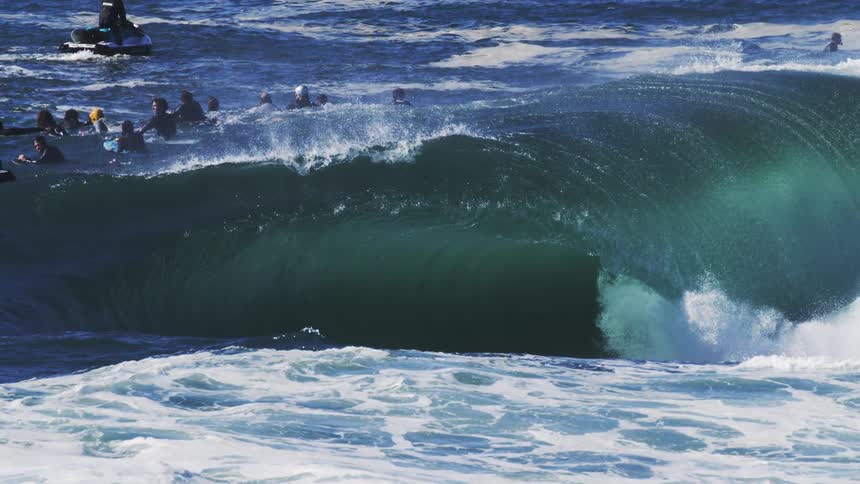 A massive turquoise wave cresting over a group of surfers waiting for their chance to ride