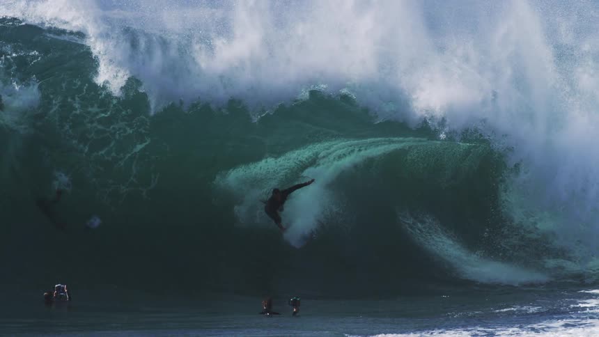 A surfer in a dark wetsuit falls from their board swallowed by the hollow of a massive surging wave
