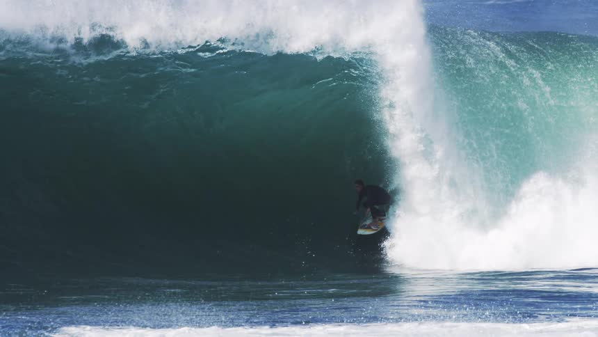 Sunlight shines upon a huge wave as it splashes and curls over a surfer