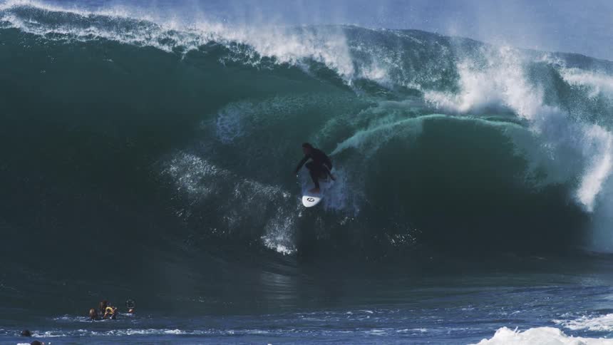 A lone surfer expertly navigates a massive barreling wave as fellow surfers watch from the water below
