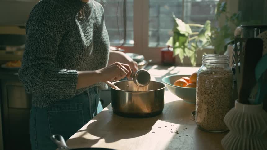 A woman serves a portion of food from a jar into a pan on the kitchen countertop and continuously stirs it as it cooks