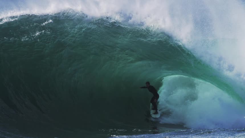 A surfer in a dark suit is engulfed by the hollow of a massive powerful wave