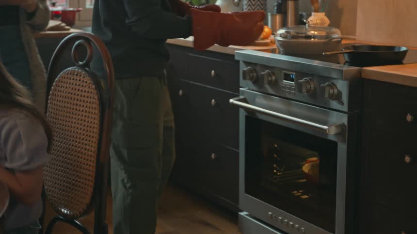 A family cooks dinner while the father pulls a pot of roast chicken out of the oven