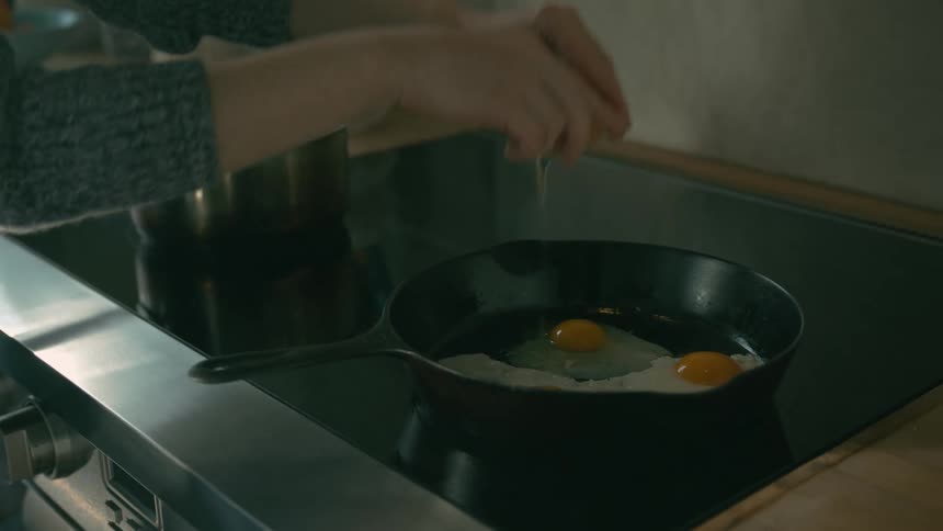 A woman is cracking eggs into a frying pan on the stove in the kitchen