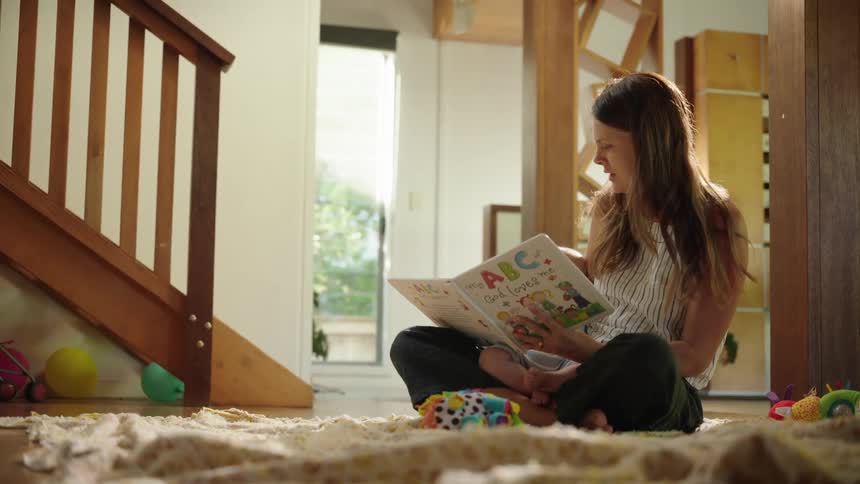 A woman sits on the living room floor holding her infant child and reading a book out loud