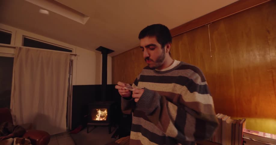 A man wearing a sweatshirt picks up a piece of paper from a bowl on a coffee table then drops it back and begins gesturing as his friends sit on couches in the living room at night with a fireplace glowing behind him