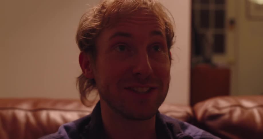 A man with light curly hair and a plaid shirt is smiling while resting his chin on his hand