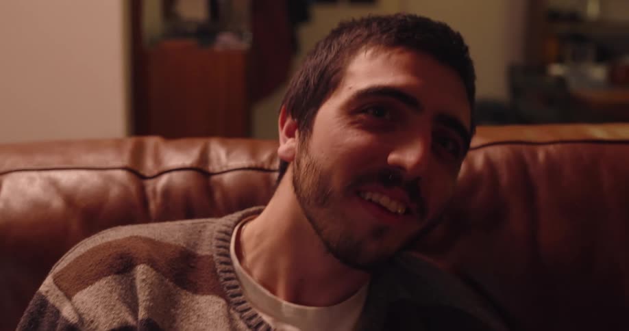 A man wearing a sweatshirt sits on a sofa laughing