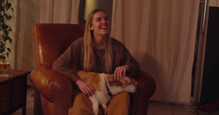 A smiling woman sits in a leather armchair with a dog on her lap celebrating with her partner winning a game of charades