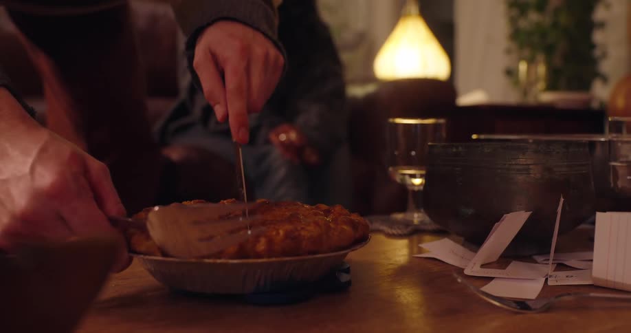 A persons hands use a fork and knife to cut into a small goldenbrown pie in a foil tin on a wooden table cluttered with papers and dishes