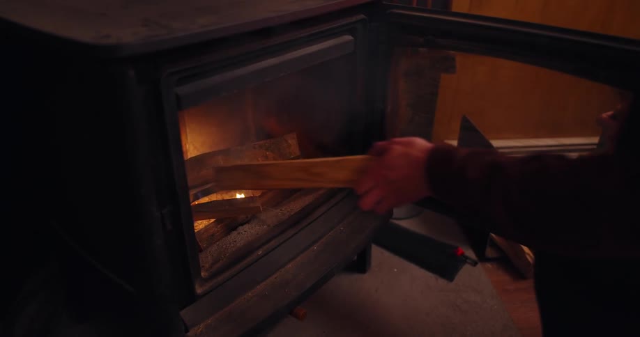 A person adds a piece of wood to a fire burning inside a black woodburning stove
