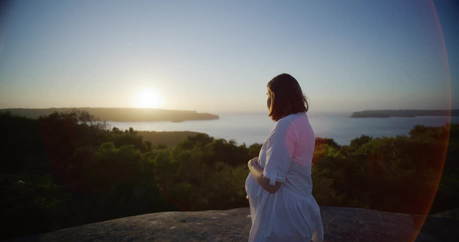 Pregnant woman in white dress on the peak of a rock with a sunrise on the horizon