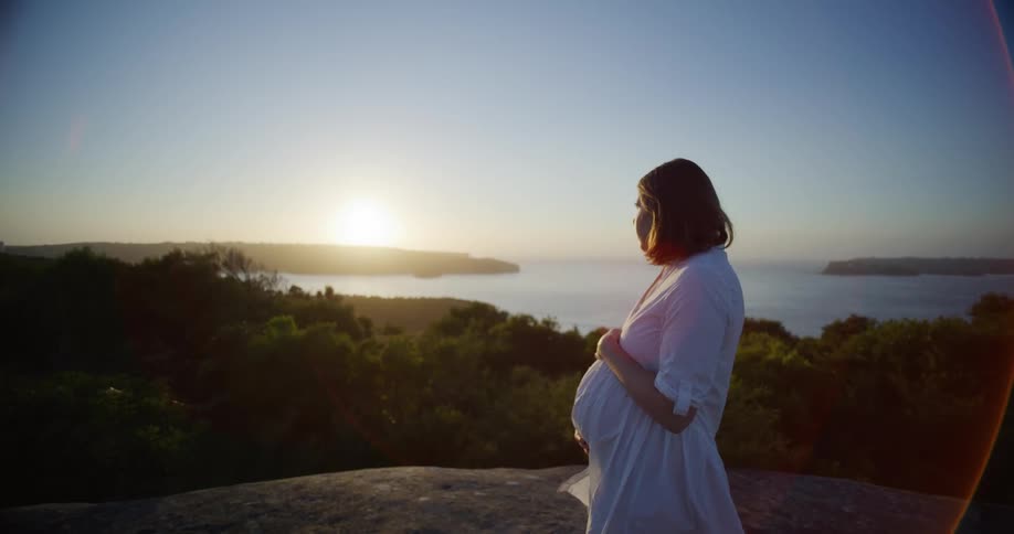 Pregnant woman in white dress on a high vantage point of a rock overlooking a lake landscape and the sun rising on the horizon
