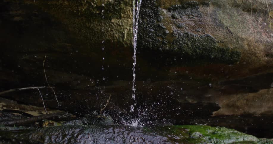 A thin stream of water drips steadily from a mosscovered rock overhang splashing onto a wet rock below and sending tiny droplets into the air