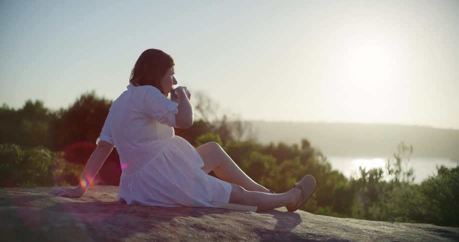 A woman in a white dress sits on a rock at sunset sipping from a cup and gazing over a serene sunlit landscape
