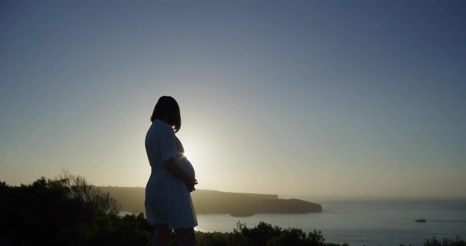 A pregnant woman stands on a cliff at sunrise silhouetted against the coastal horizon
