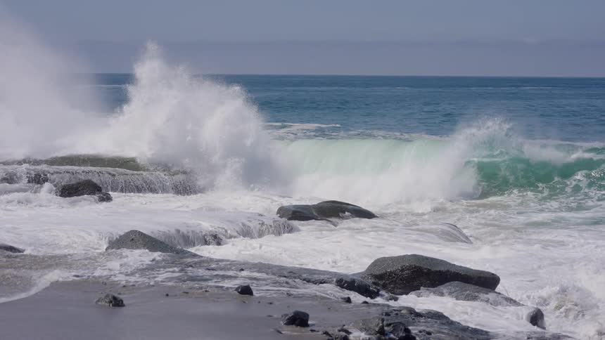 Powerful ocean waves crash dramatically against a rocky shoreline sending plumes of white sea spray into the air under a hazy sky
