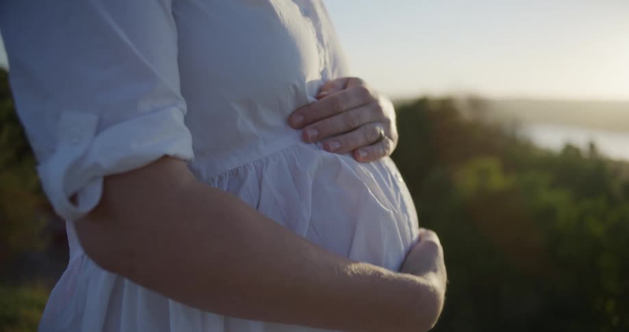 A pregnant person in a white dress gently cradles their baby bump against a softly blurred natural backdrop at golden hour