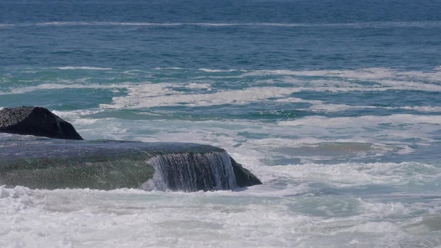 Ocean water continuously flows over a flat dark rock creating a small cascade into foamy white surf with the blue and white choppy sea visible beyond