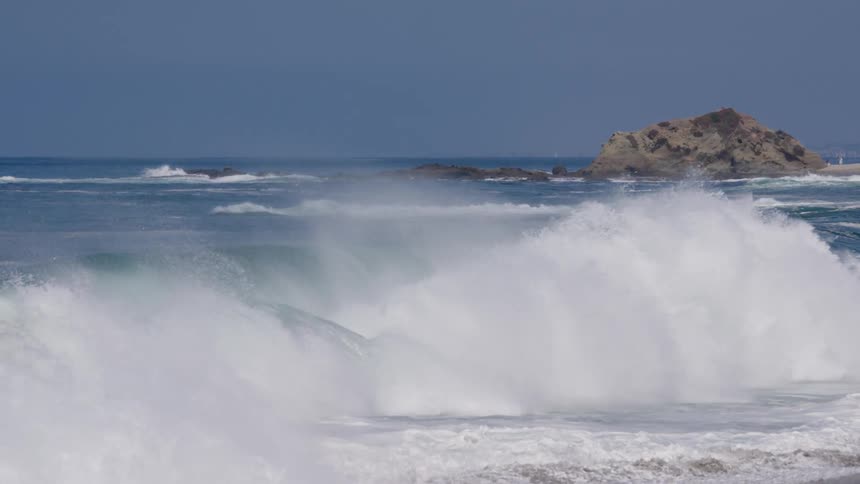 Large ocean waves with green translucent crests break powerfully sending up white sea spray as they roll towards the shore with a rocky outcrop visible in the background under a clear blue sky