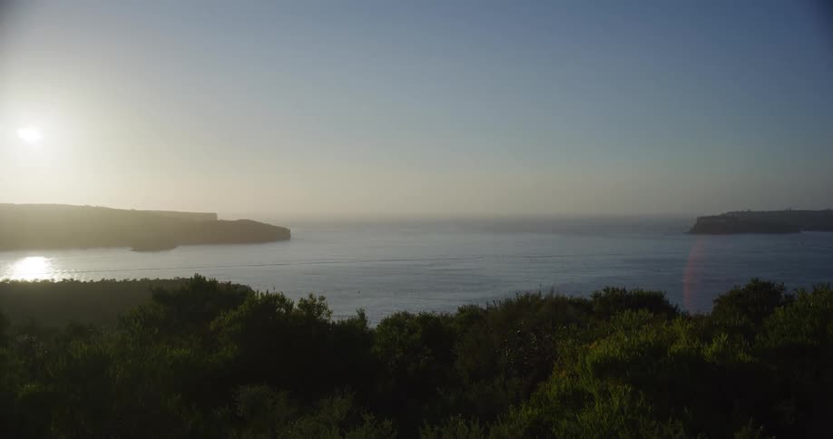 A hazy sunrise or sunset casts a bright glare over a calm ocean inlet viewed from above a silhouetted tree line with distant headlands framing the water