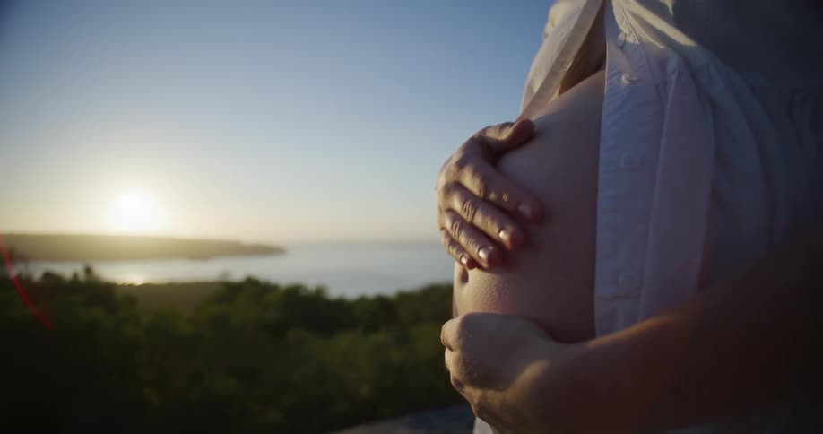 Pregnant woman rubs her belly while standing close to the sea at sunset