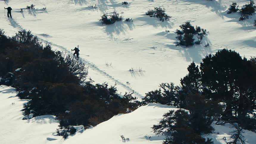Two people trek through a snowy landscape following a winding path surrounded by bushes and sparse trees