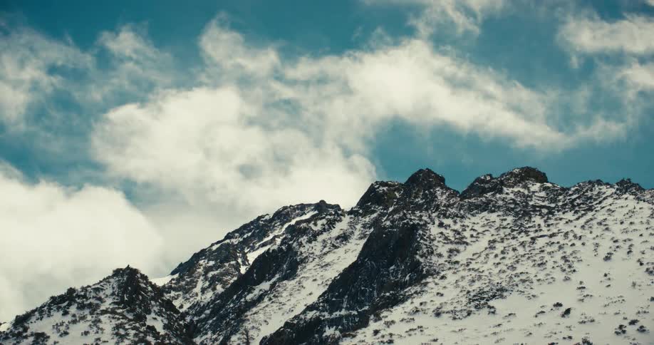 Jagged snowdusted mountain peaks rise against a vibrant blue sky dotted with wispy white clouds