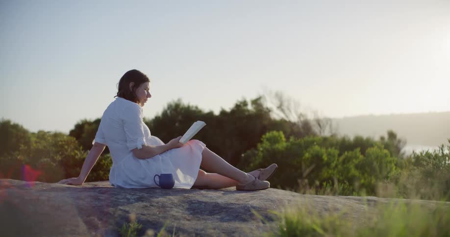 Pregnant woman sitting on a rock outdoors reading a book