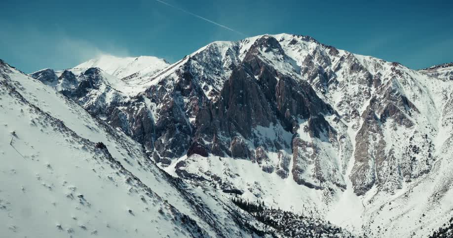 An aerial shot reveals a majestic snowcovered mountain range with rugged rocky peaks reaching towards a clear blue sky with a distant jet contrail visible