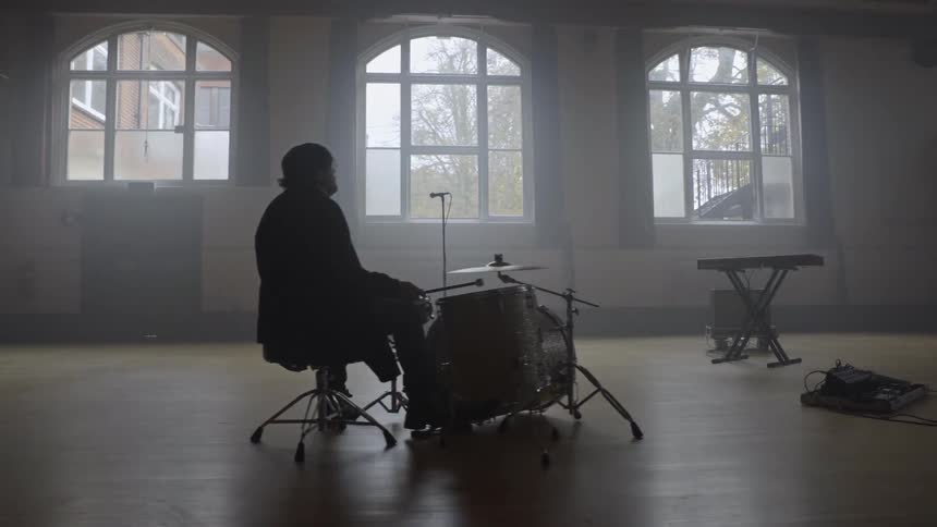A lone drummer sits behind a drum kit in a spacious empty hall illuminated by natural light from tall windows
