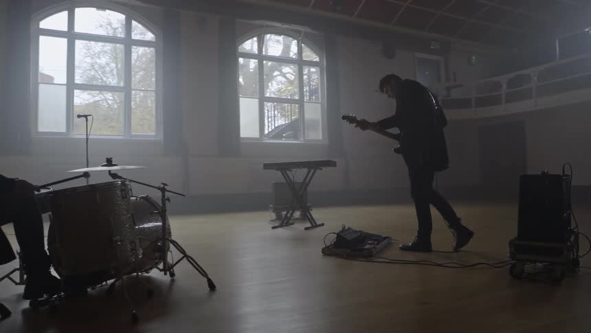 In a dimly lit spacious hall two musicians perform together one playing the drums while the other strums a guitar