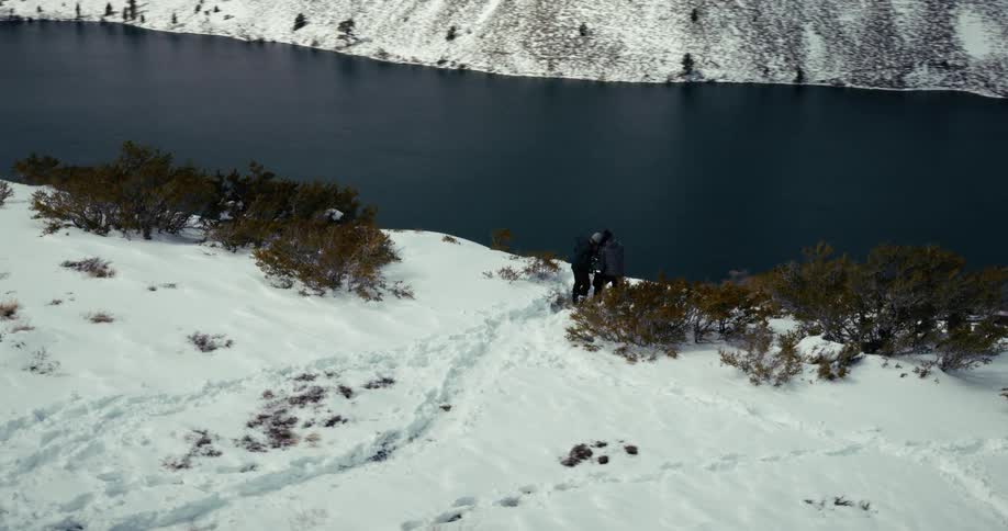 Three people on the peak of a snowy mountain overlooking a lake in the background