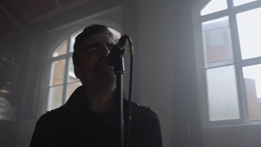 A male vocalist sings passionately into a microphone as the camera orbits him during a band rehearsal in a dimly lit studio