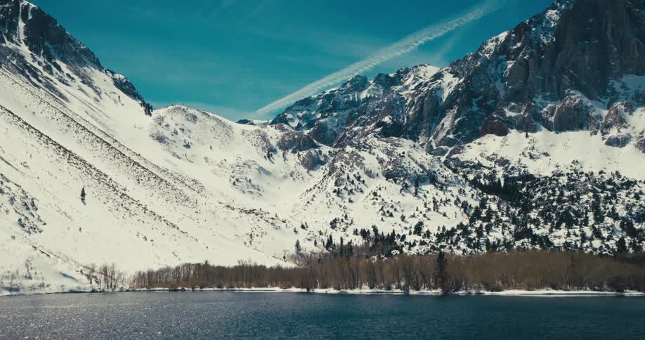 A serene landscape features a placid lake and snowcapped mountains in the distance all under a sky filled with soft clouds